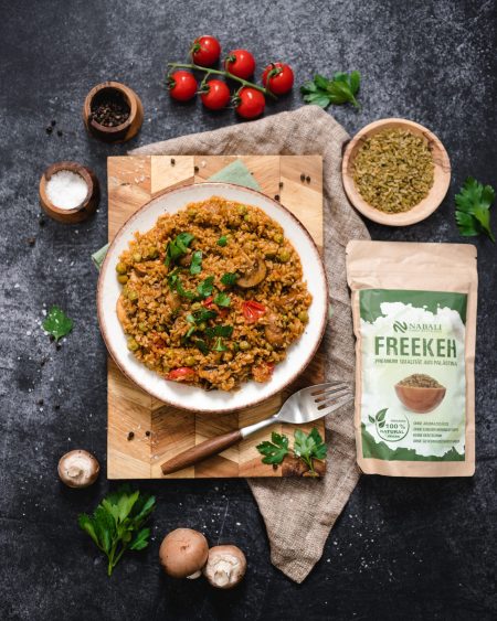 Ein Teller Freekeh mit Gemüse und Kräutern, umgeben von frischen Zutaten und einer Packung Freekeh.