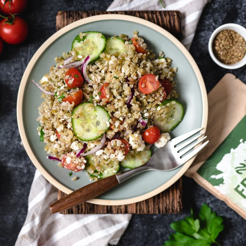 Frischer Freekeh Salat mit Gurken und Tomaten Ein frischer Freekeh-Salat mit Gurkenscheiben, Kirschtomaten, roten Zwiebeln und Feta-Käse, auf einem rustikalen Holzbrett serviert.